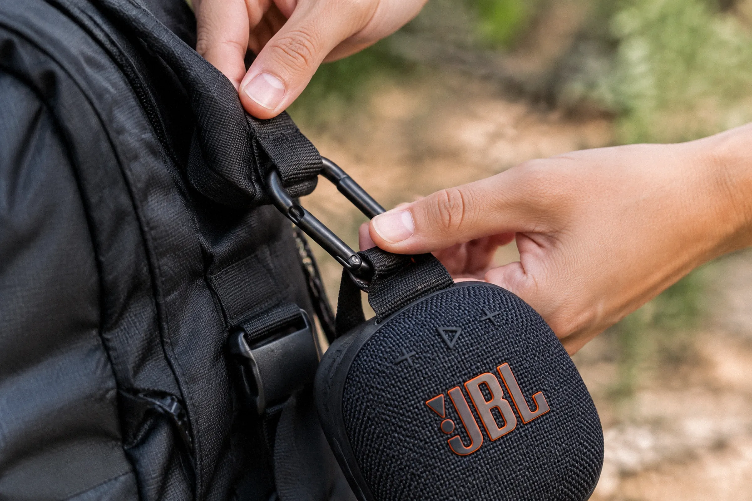 Close nas mãos encaixando o mosquetão de uma caixa de som na alça de uma mochila, com o ponto de fixação totalmente visível.